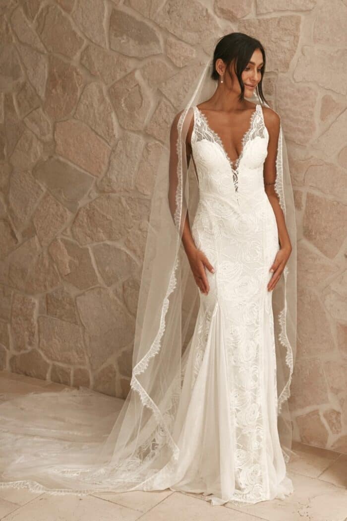 A model wears a lace wedding dress with plunge v neckline and veil against a rustic setting.