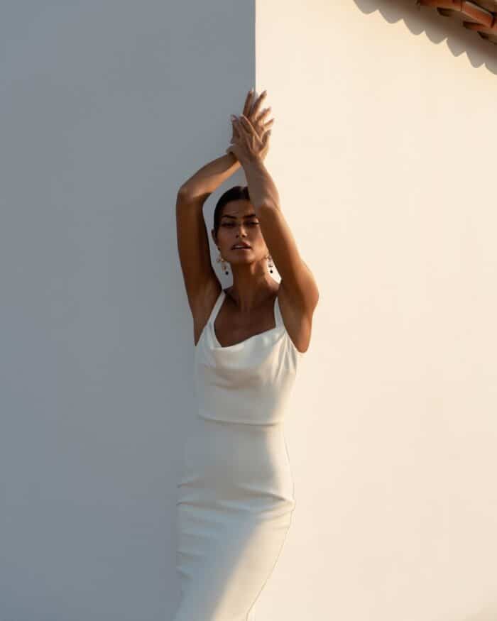 Model wears a minimalist sleeveless square neck wedding dress with arms raised.
